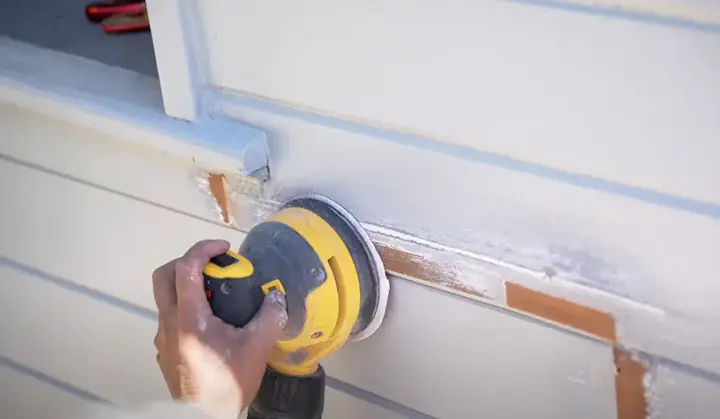 Hand using a yellow power sander on an exterior wall near a window, surrounded by white dust and wooden board detailing. Hand using a yellow power sander on an exterior wall near a window, surrounded by white dust and wooden board detailing.