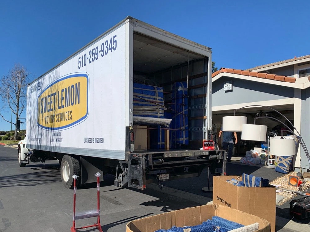 In this image, two professional movers from Sweet Lemon Moving Services are shown carefully carrying a large, wrapped sofa down a staircase inside a residential home. Both movers are wearing work uniforms and gloves, ensuring the furniture is securely handled to prevent damage. The staircase is narrow, and the movers are coordinating closely to navigate the bulky item through the confined space. This demonstrates the company's commitment to careful, efficient, and safe moving practices, making the relocation process smooth for their clients. In this image, two professional movers from Sweet Lemon Moving Services are shown carefully carrying a large, wrapped sofa down a staircase inside a residential home. Both movers are wearing work uniforms and gloves, ensuring the furniture is securely handled to prevent damage. The staircase is narrow, and the movers are coordinating closely to navigate the bulky item through the confined space. This demonstrates the company's commitment to careful, efficient, and safe moving practices, making the relocation process smooth for their clients.