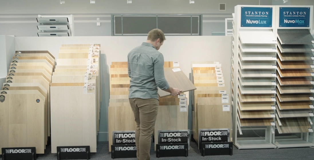 Person browsing through various flooring samples in a showroom, with multiple display racks of wood and laminate options visible. Person browsing through various flooring samples in a showroom, with multiple display racks of wood and laminate options visible.
