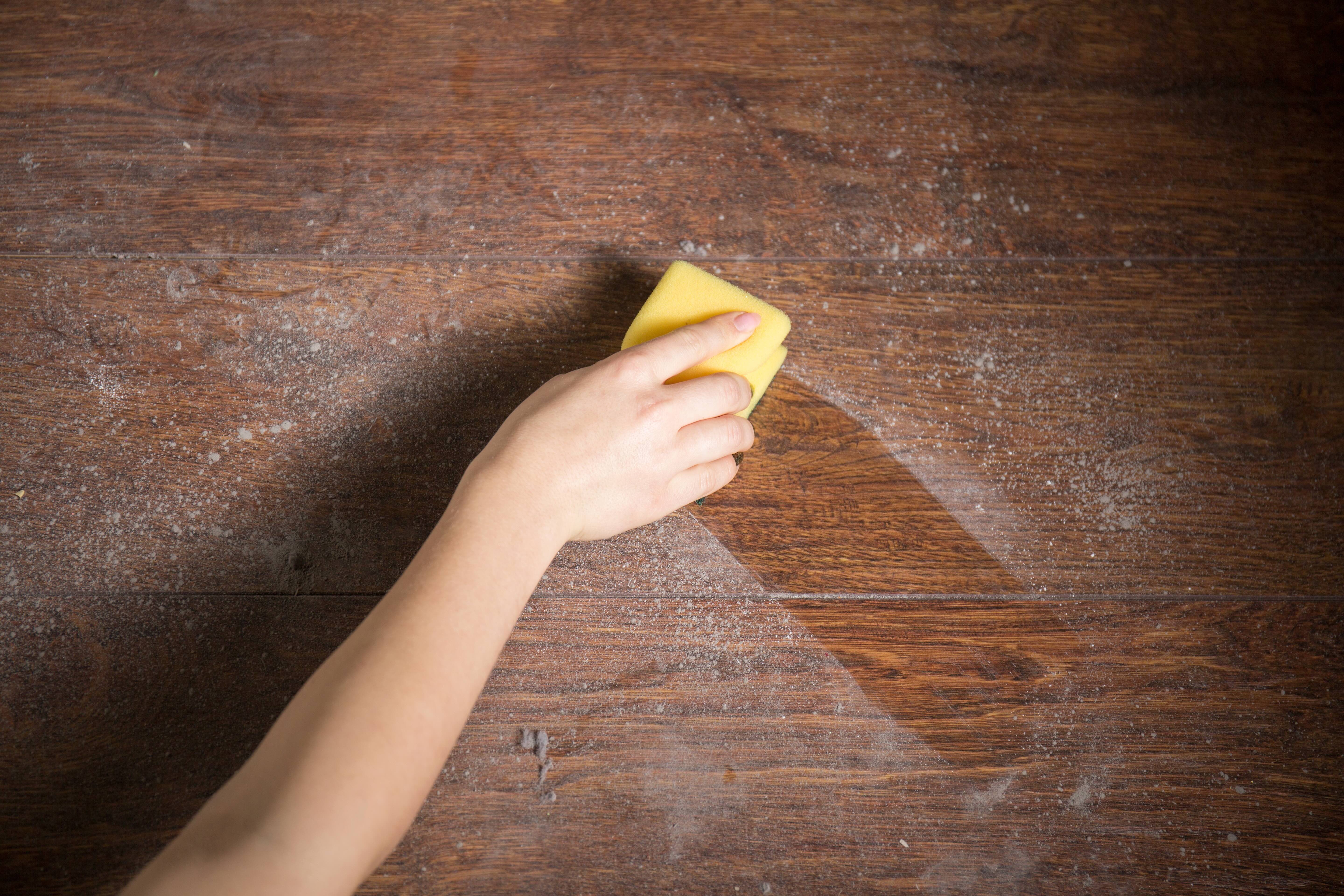 A person wipes a thick layer of dust from a wooden surface with a yellow sponge.