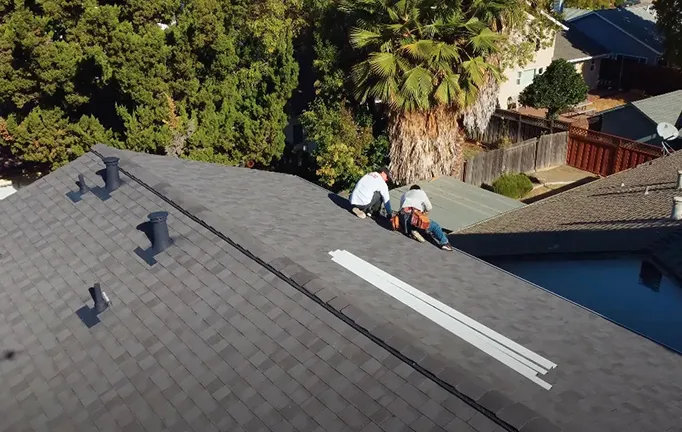  Two workers in white shirts adjust shingles on a residential rooftop surrounded by trees and neighboring houses. Long strips of material lie beside them.