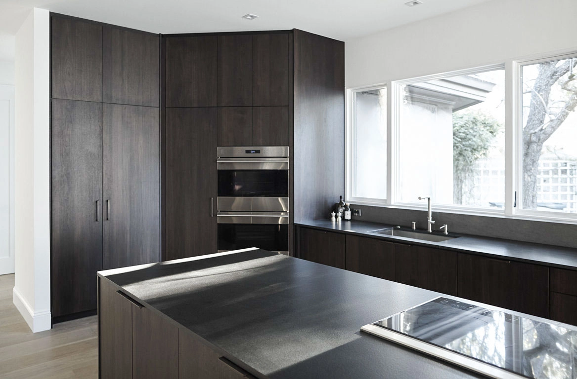  Modern kitchen featuring dark wood cabinets, built-in oven, sleek countertops, and large windows providing natural light.