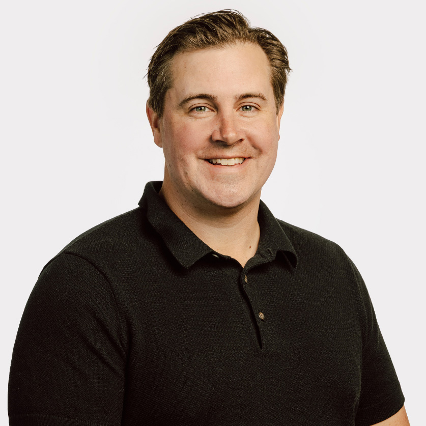 Headshot of Craig Baldwin wearing a black collared shirt against a light gray background.