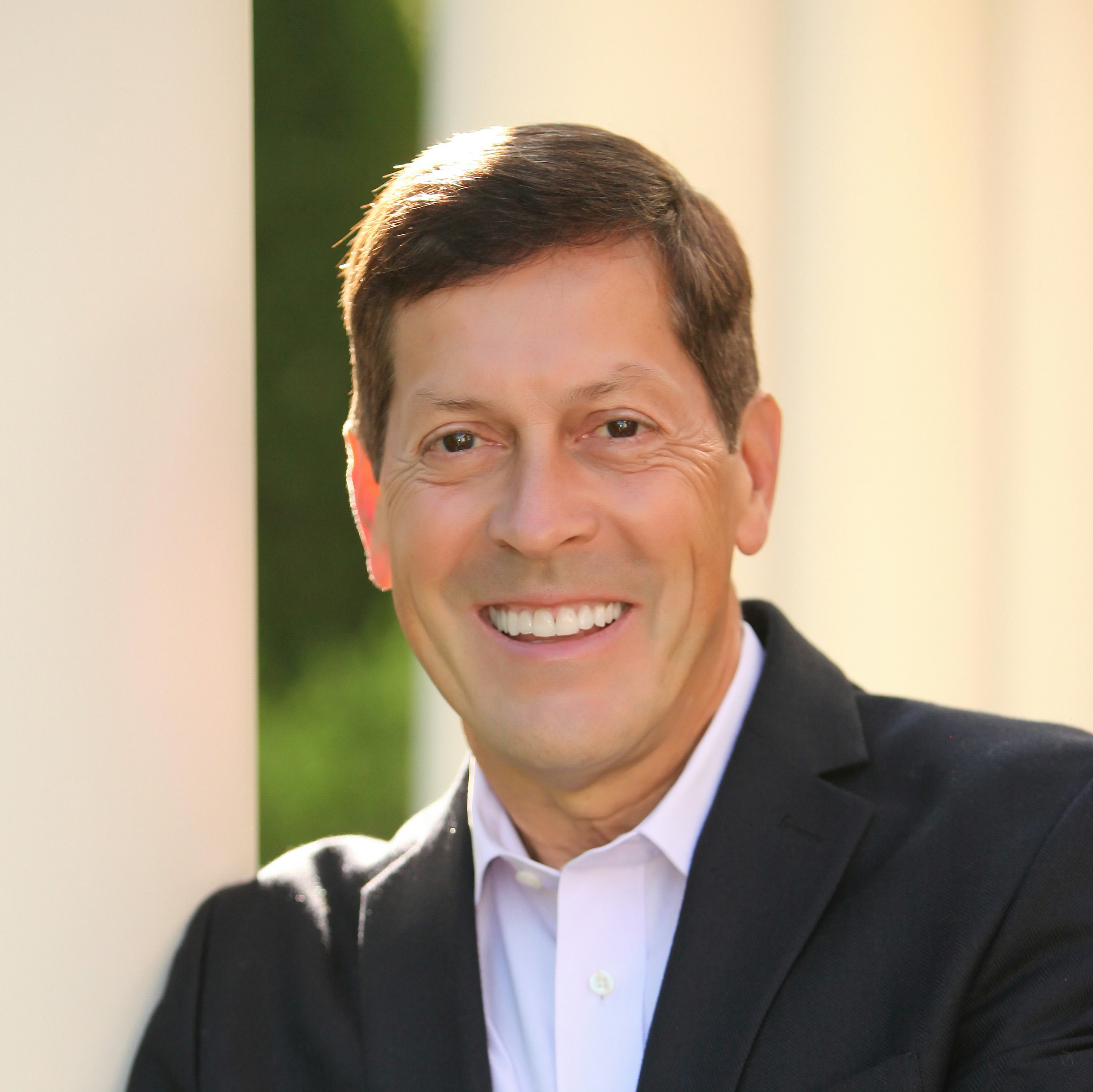 Headshot of Ric Dillon in a dark blazer and light collared shirt, with a softly blurred outdoor background featuring columns and greenery.
