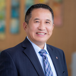 Headshot of Henry Xiang wearing a pale blue button up shirt, blue plaid tie and dark colored suit jacket