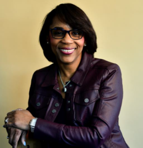Headshot of Mae Smitherman-Smith in glasses and a burgundy leather jacket, seated against a plain beige background.