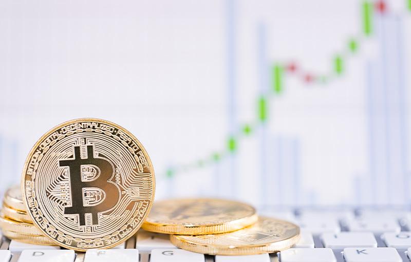 Gold color coins with Bitcoin logo siting on top of a keyboard with blurry chart in the background