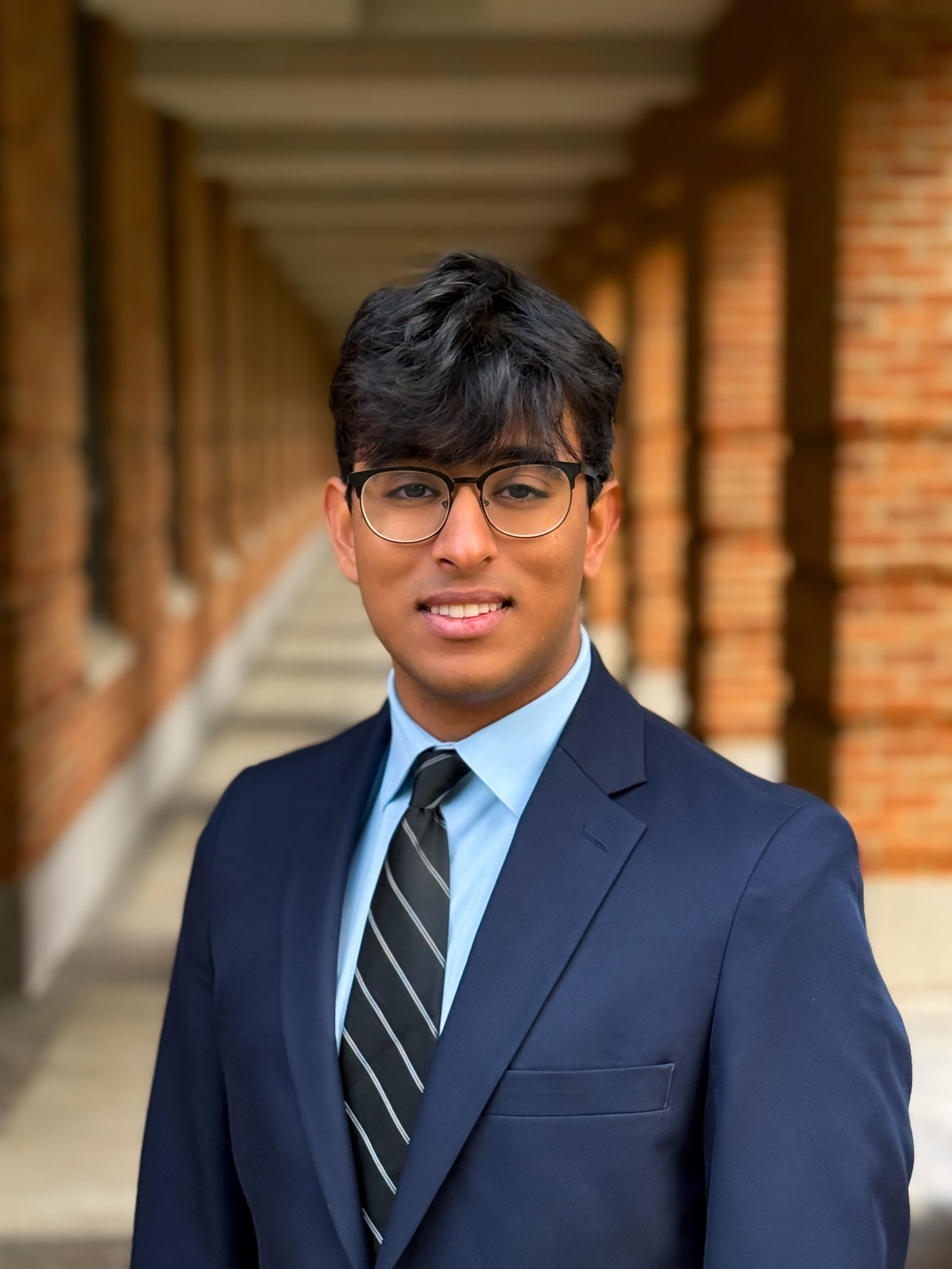 man with glasses smiling at camera with navy blue suit, baby blue dress shirt, and black tie