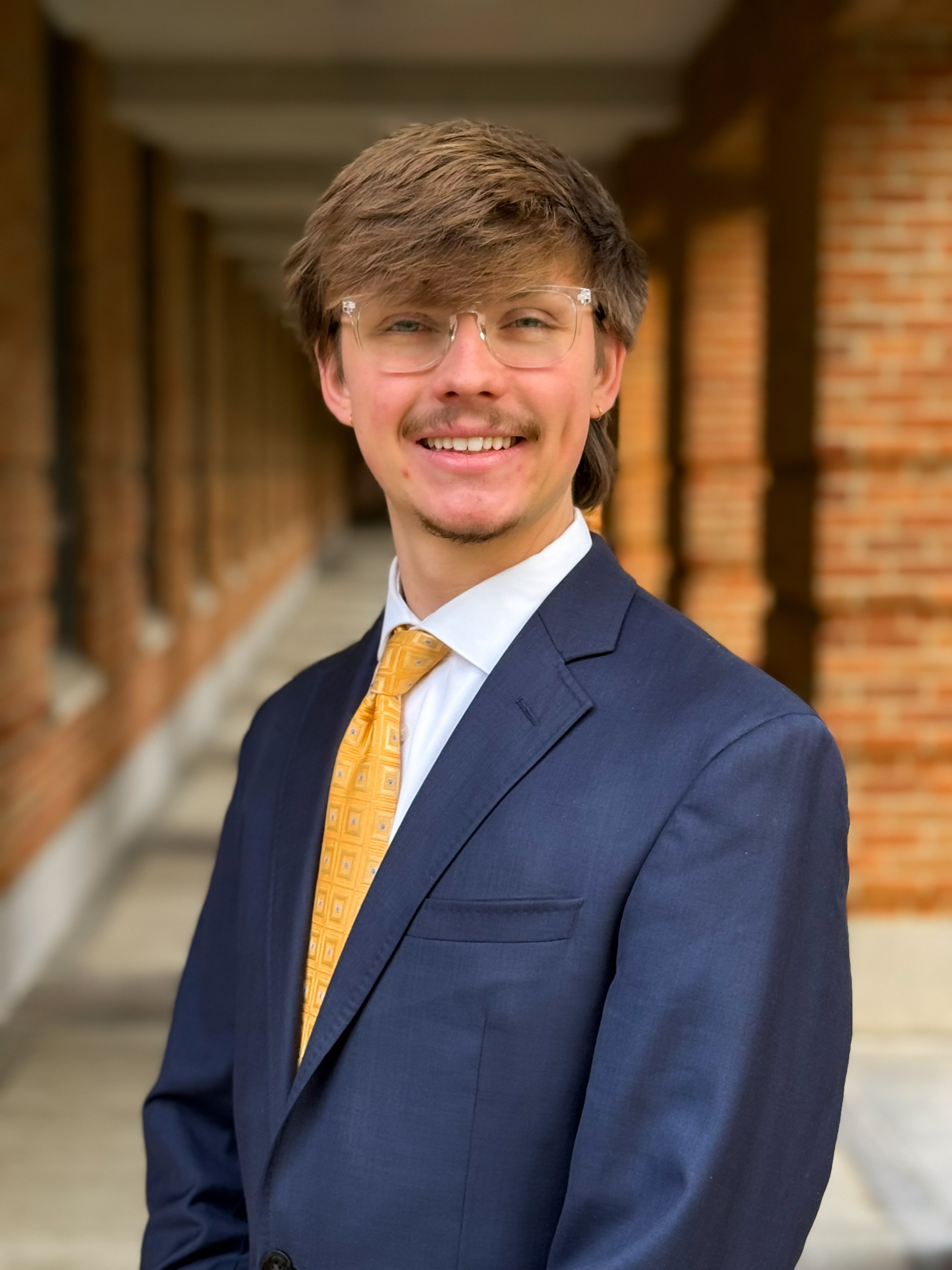 man with glasses smiling at camera with blue suit and yellow/gold tie