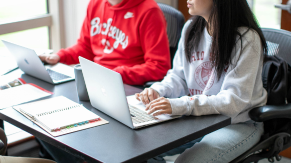 Two students sitting on a desk working on computers.