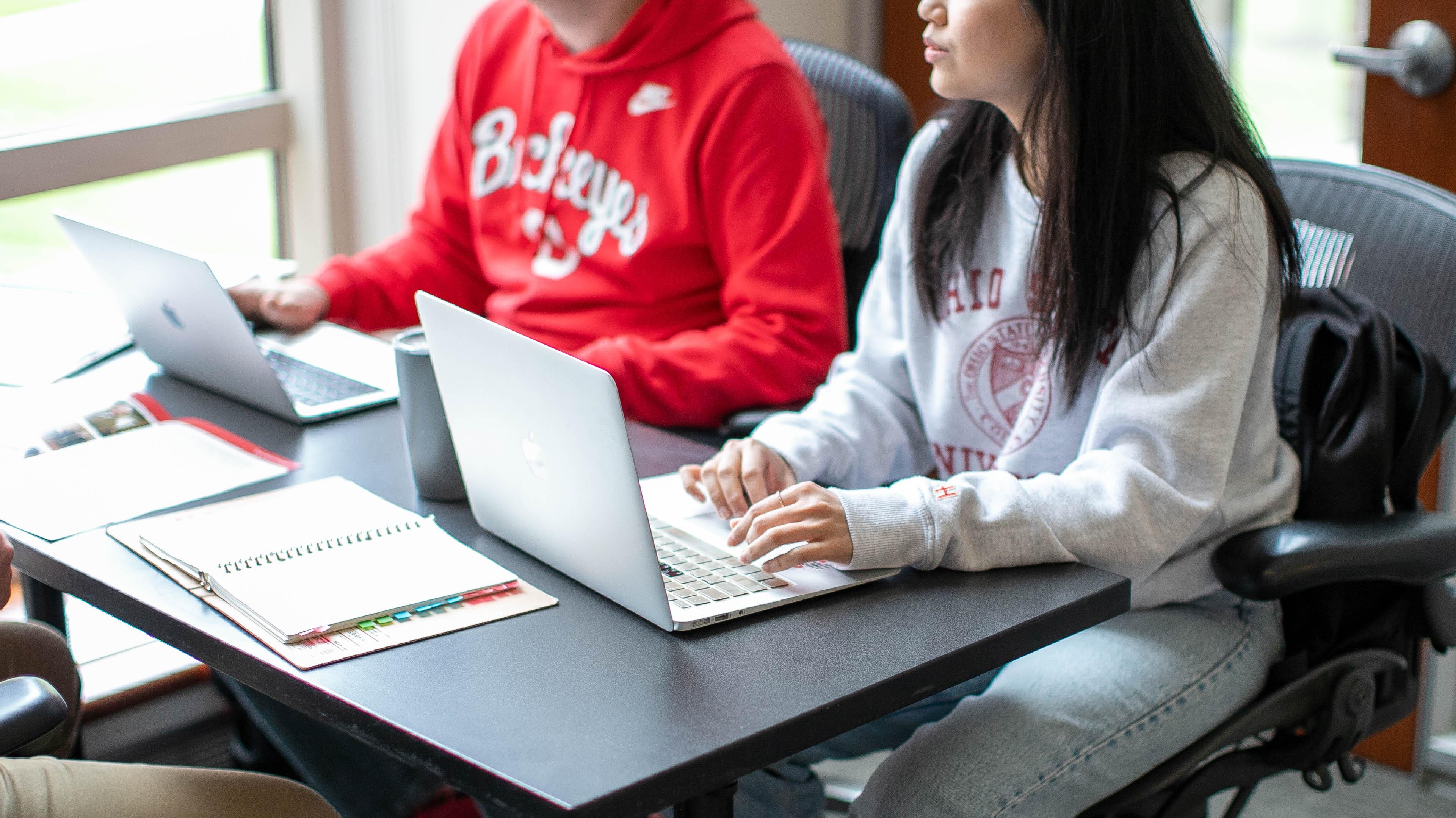 Two students sitting on a desk working on computers.
