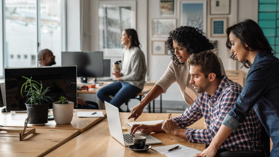 3 people on a business team having a discussion at a desk with 2 people having another discussion in the background