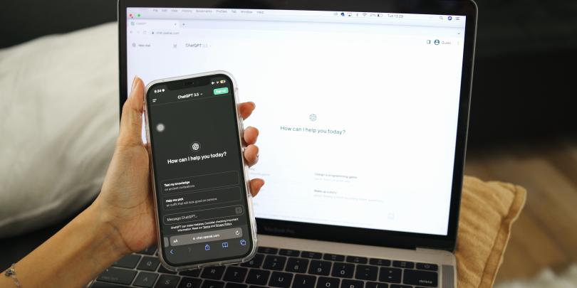 A hand holding a phone open in the ChatGPT app in front of a laptop