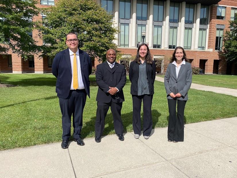 A photograph of four individuals in a line in front of a college. 