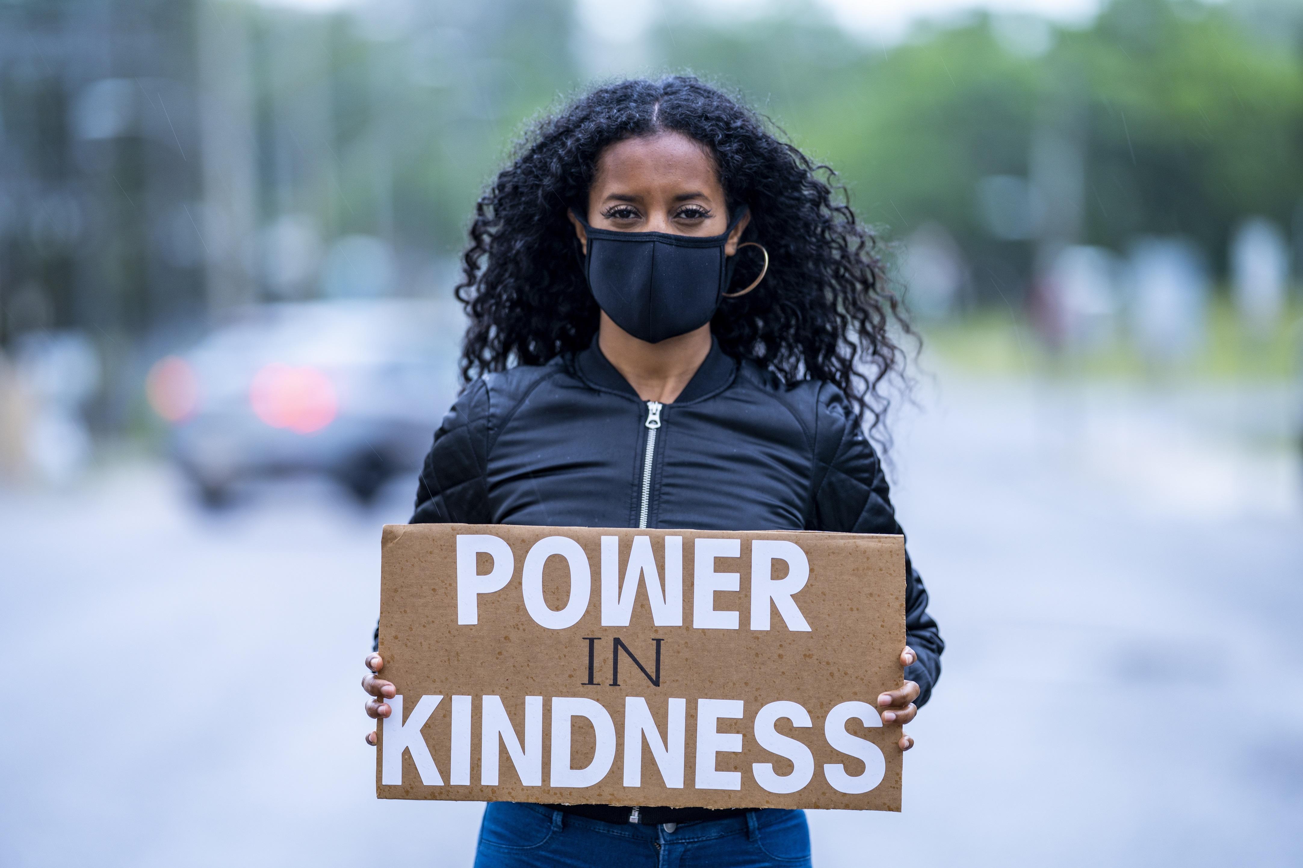 Woman in mask holding sign that says Power in Kindness