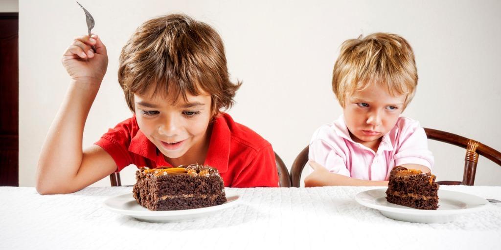 two children eating cake; one looks excited and the other grumpy
