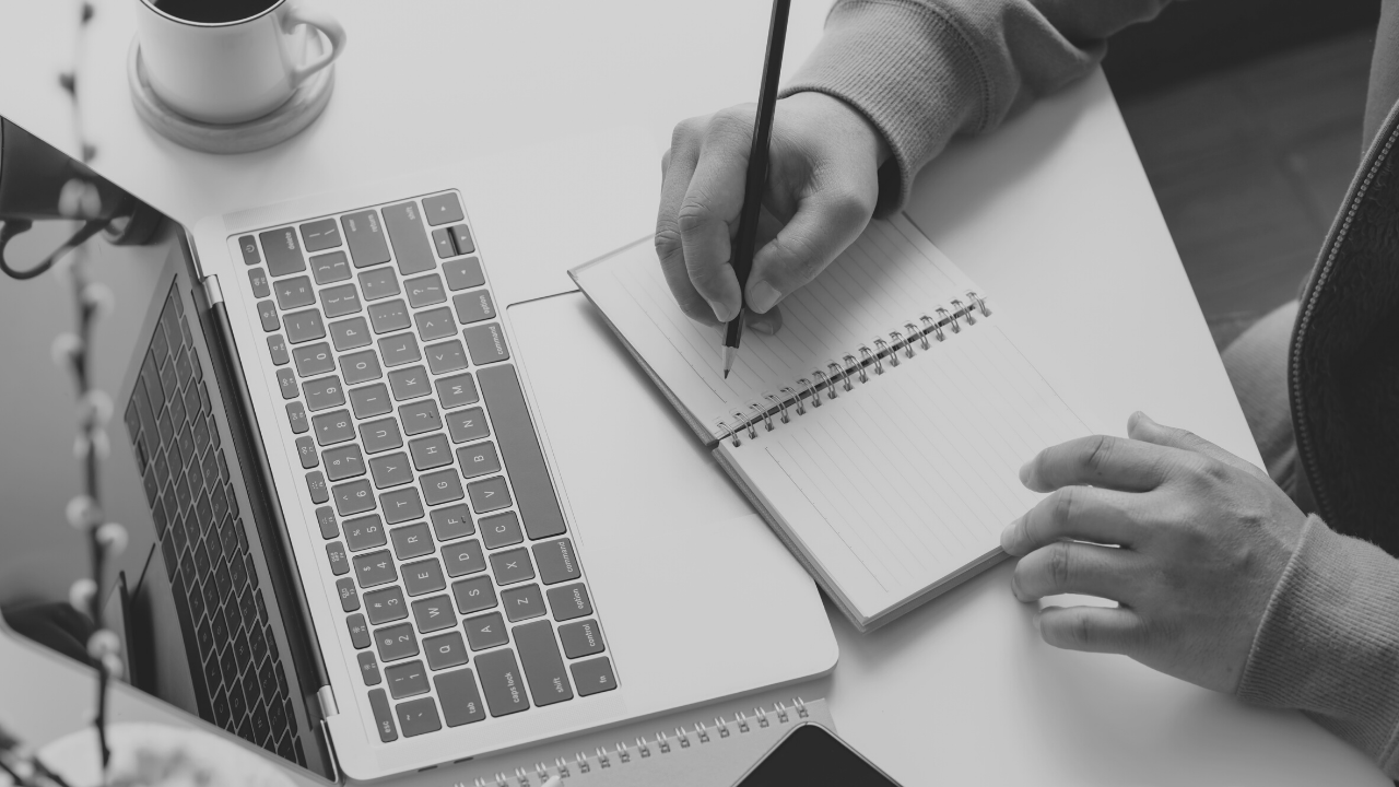 Black and white image of someone taking notes in a notebook next to their open laptop