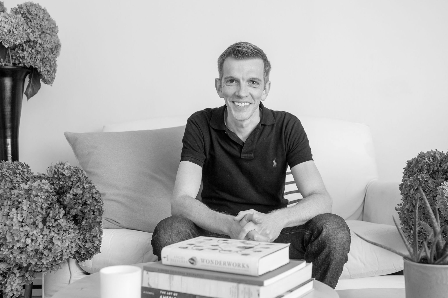 Black and white image of Angus Fletcher (author) sitting on a couch in front of a pile of books.