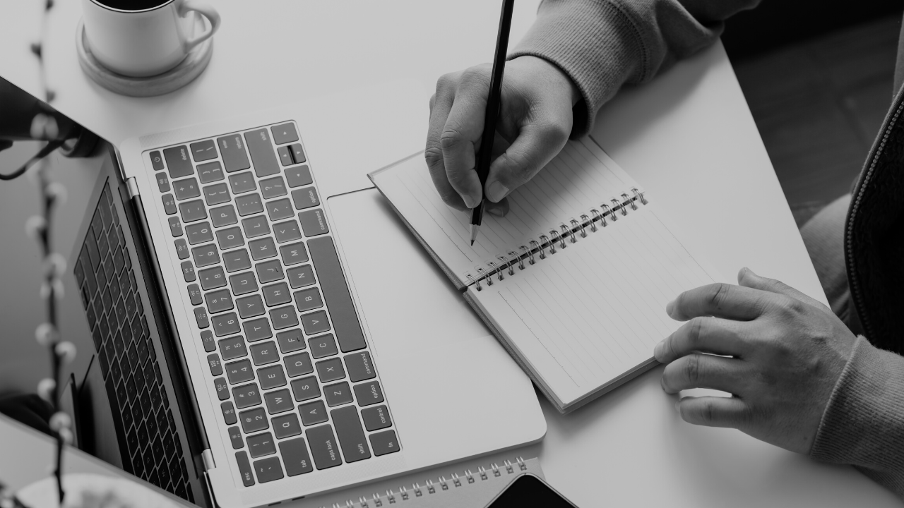 Black and white image of hands writing in a notebook with laptop open on desk