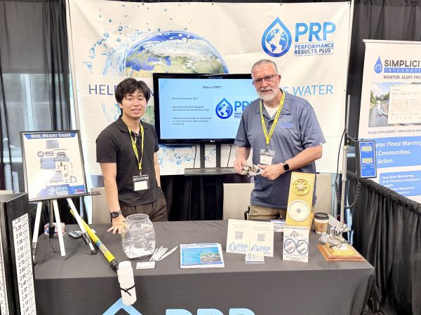 Two men standing in a trade show booth with company sign and products displayed on tabel.
