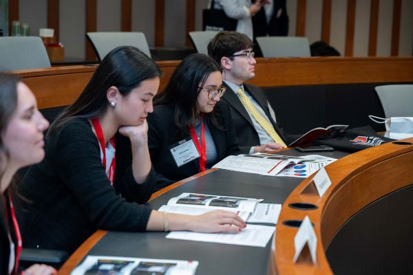 Four students in business attire sit along a curved classroom looking over information.