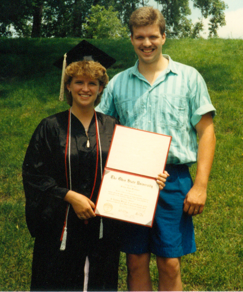 Melinda Whittington on her graduation day in 1989, holding her diploma and standing next to her husband Steven outside with trees in the background.