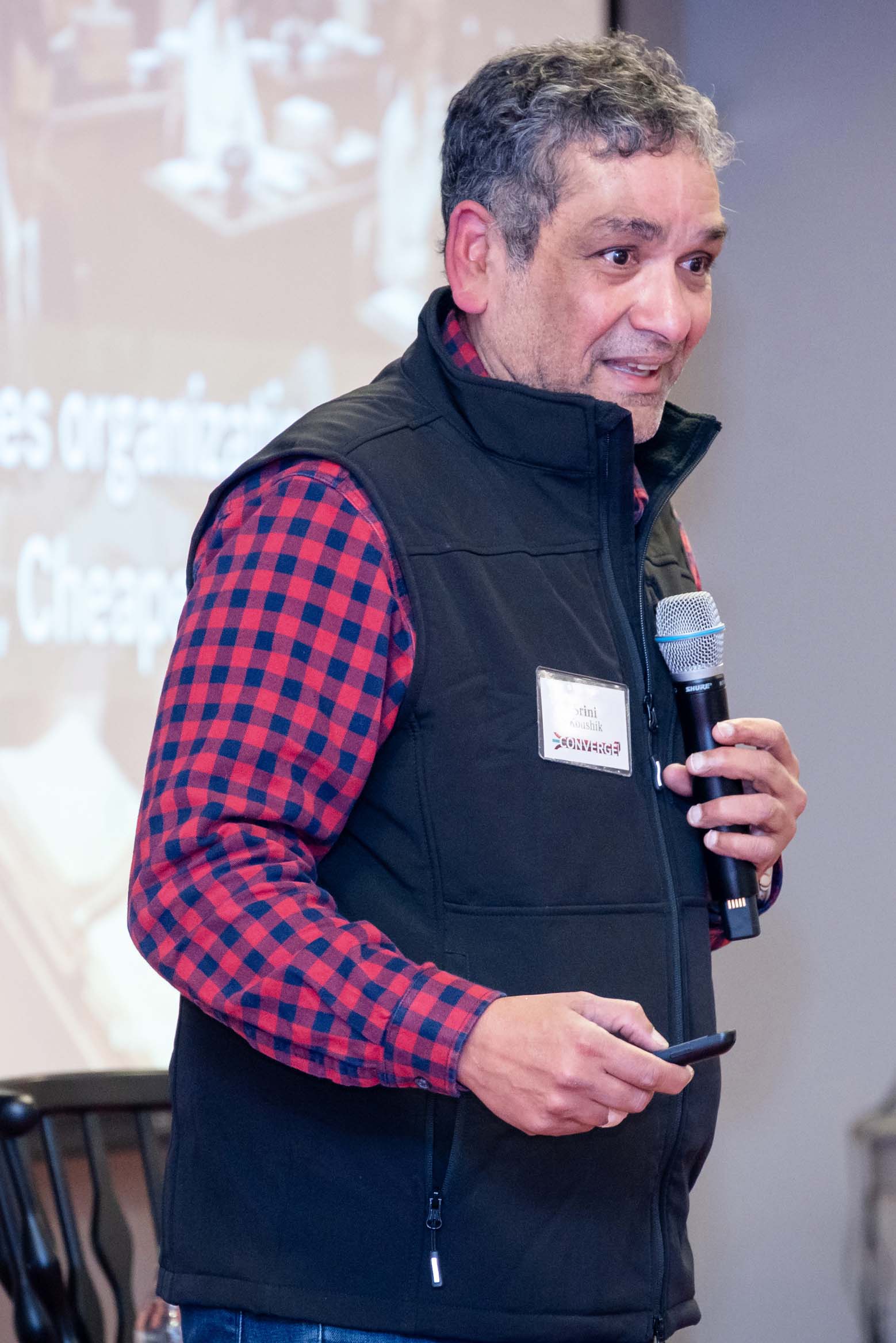 Srini Kroushik is standing at the front of a room with a screen behind him and he is holding a power point clicker. 