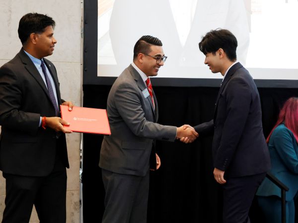 Three men in suits on stage, one holding certificate and one shaking the third's hand