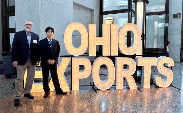 Two men in suits standing by a lit up sign that says Ohio Exports