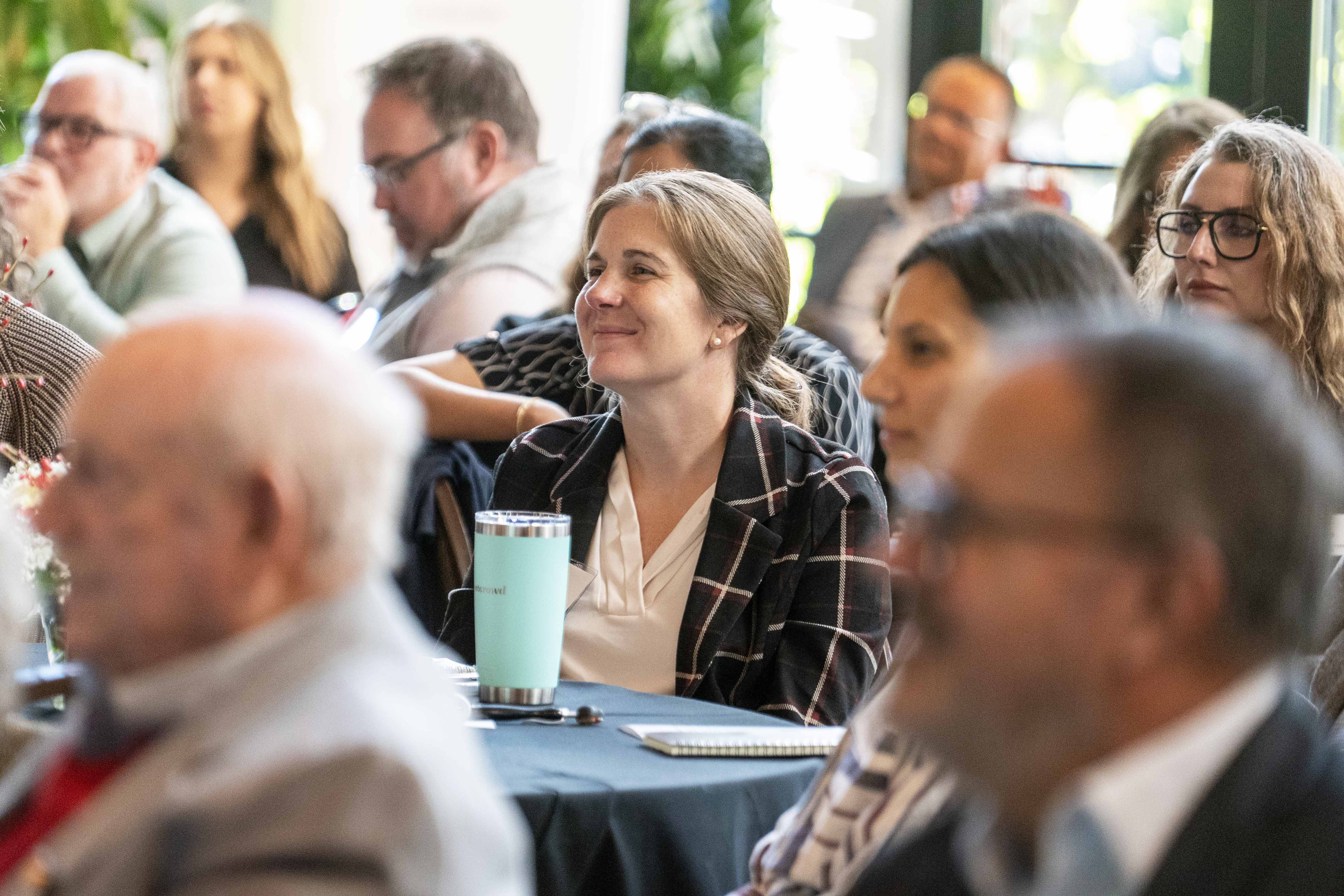 Alex Anthony in a plaid jacket and white blouse is seated at a table with other symposium attendees.