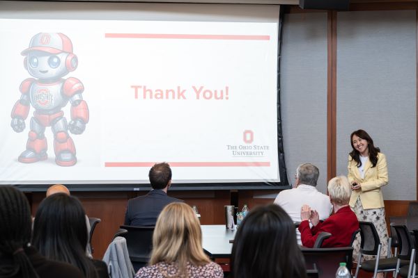 Smiling woman in dress and yellow coat next to screen displaying thank you with a robot dressed in scarlet and gray