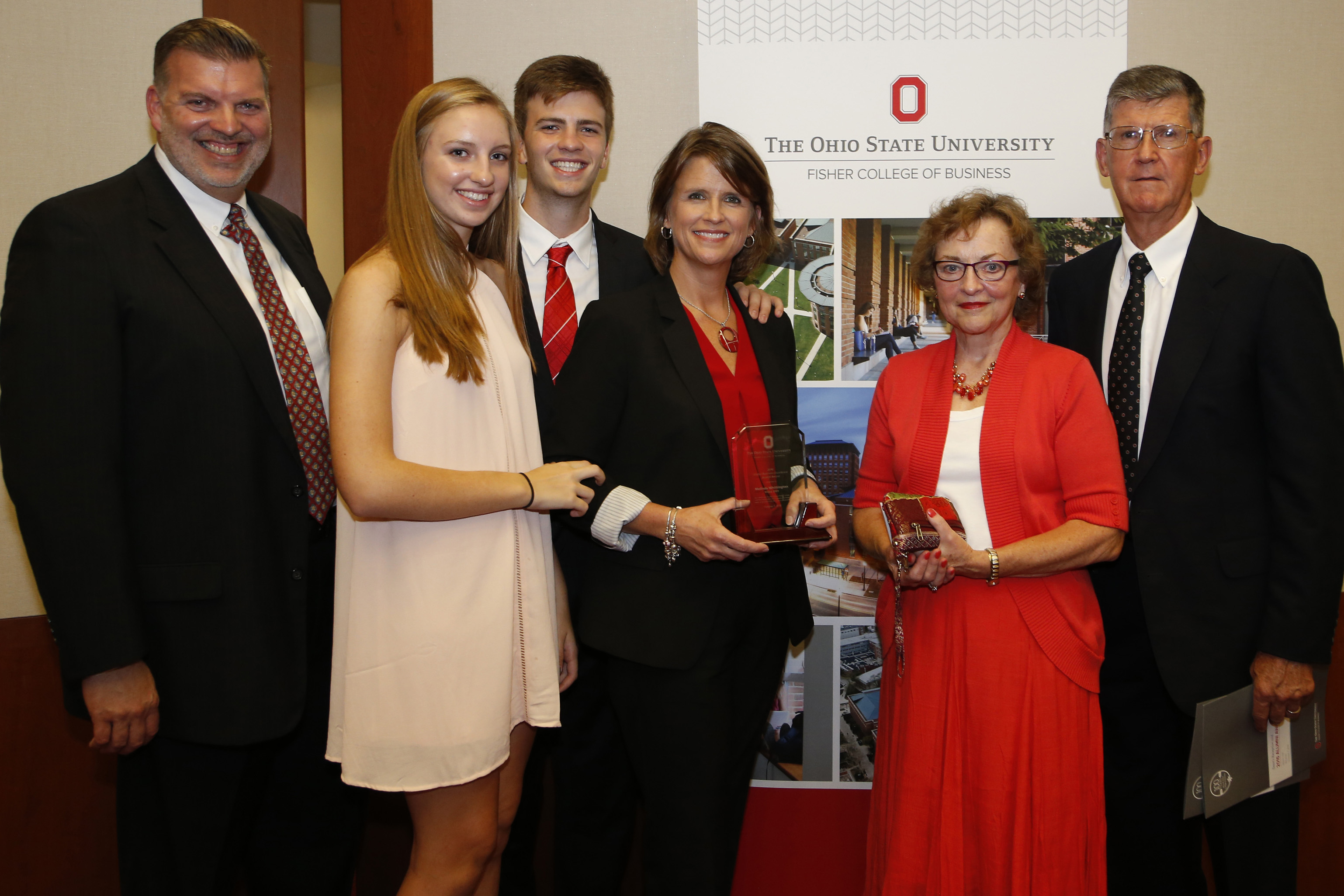 Melinda Whittington in a 2016 photo is standing in the center in a dark suit and red blouse, with her husband and two adult children on the left and her parents to the right. She is holding the Global Business Excellence Award that she received during the Alumni Awards in 2016.