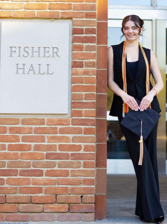 Angelino Laurino, dressed in a black pant suit, is leaning against an exterior brick wall and next to a concrete building marker that reads Fisher Hall. She is holding her graduation cap and her honors cords are draped around her neck.