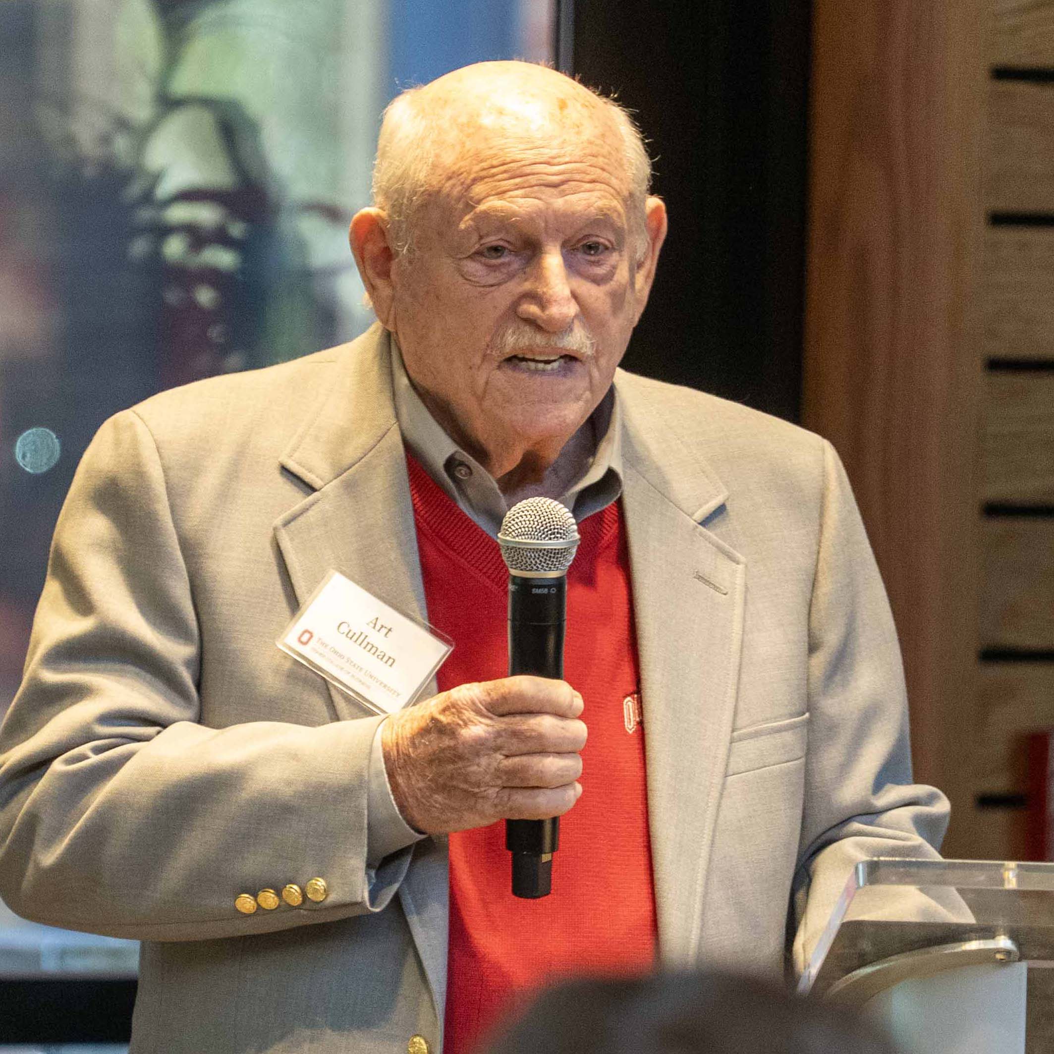 Art Cullman Jr. wearing a red Ohio State sweater under a light tan jacket and holding a microphone.