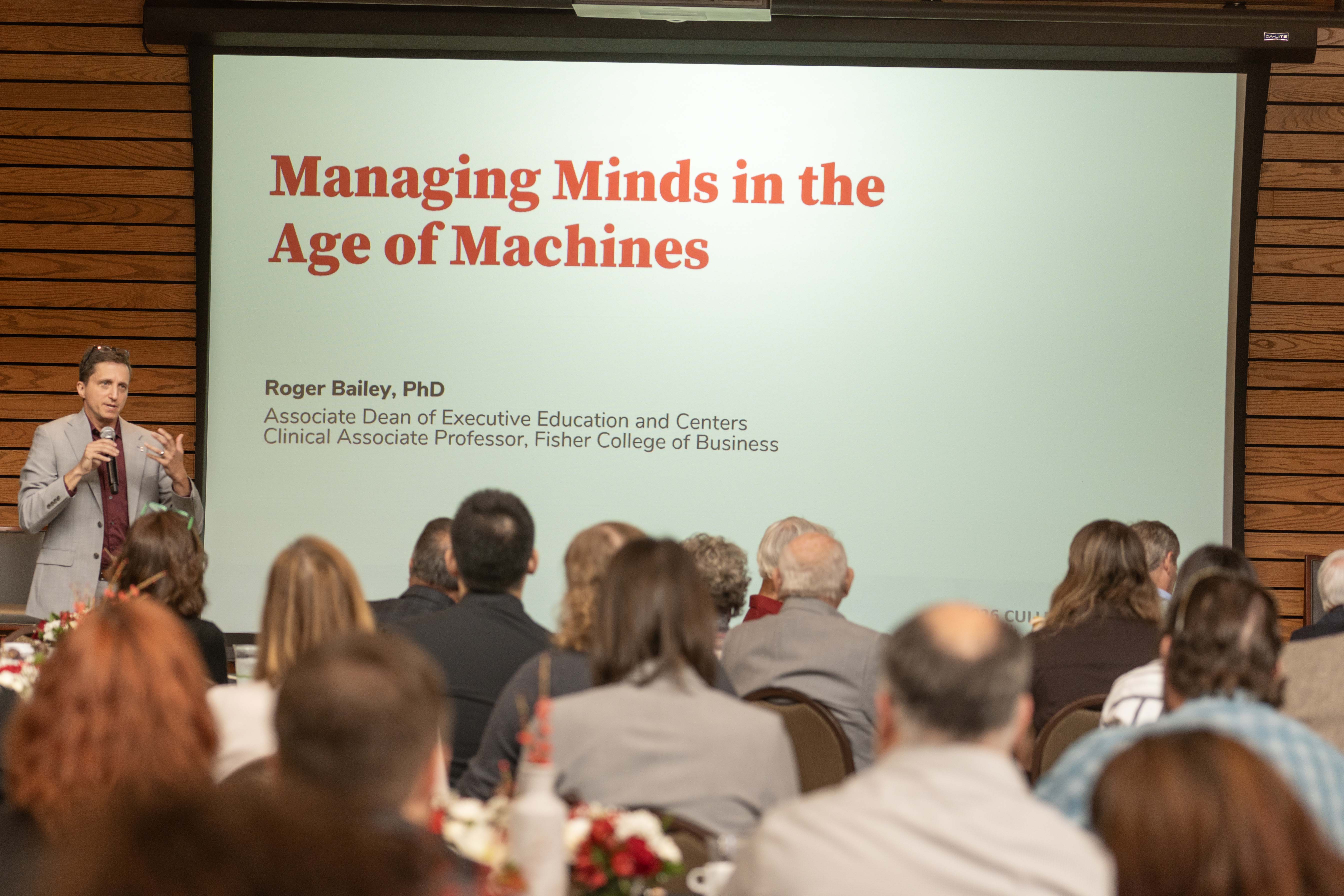 Roger Bailey stands at the front of room filled seated at tables and beside a screen that reads "Managing Minds in the Age of Machines."