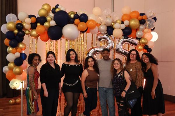 Group of 8 individuals under an orange, white and gold balloon arch with a 25 balloon