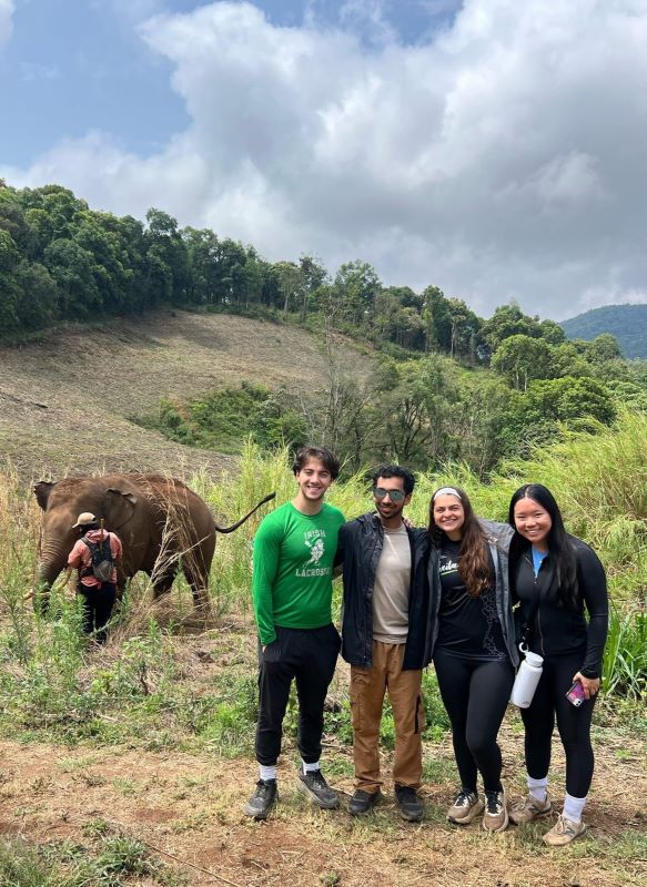 Four students in casual attire pose on a hill with an elephant and local resident in the background