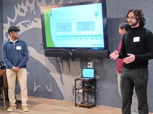 Three male students stand in front of room pointing out details on their projected Power Point presenationa