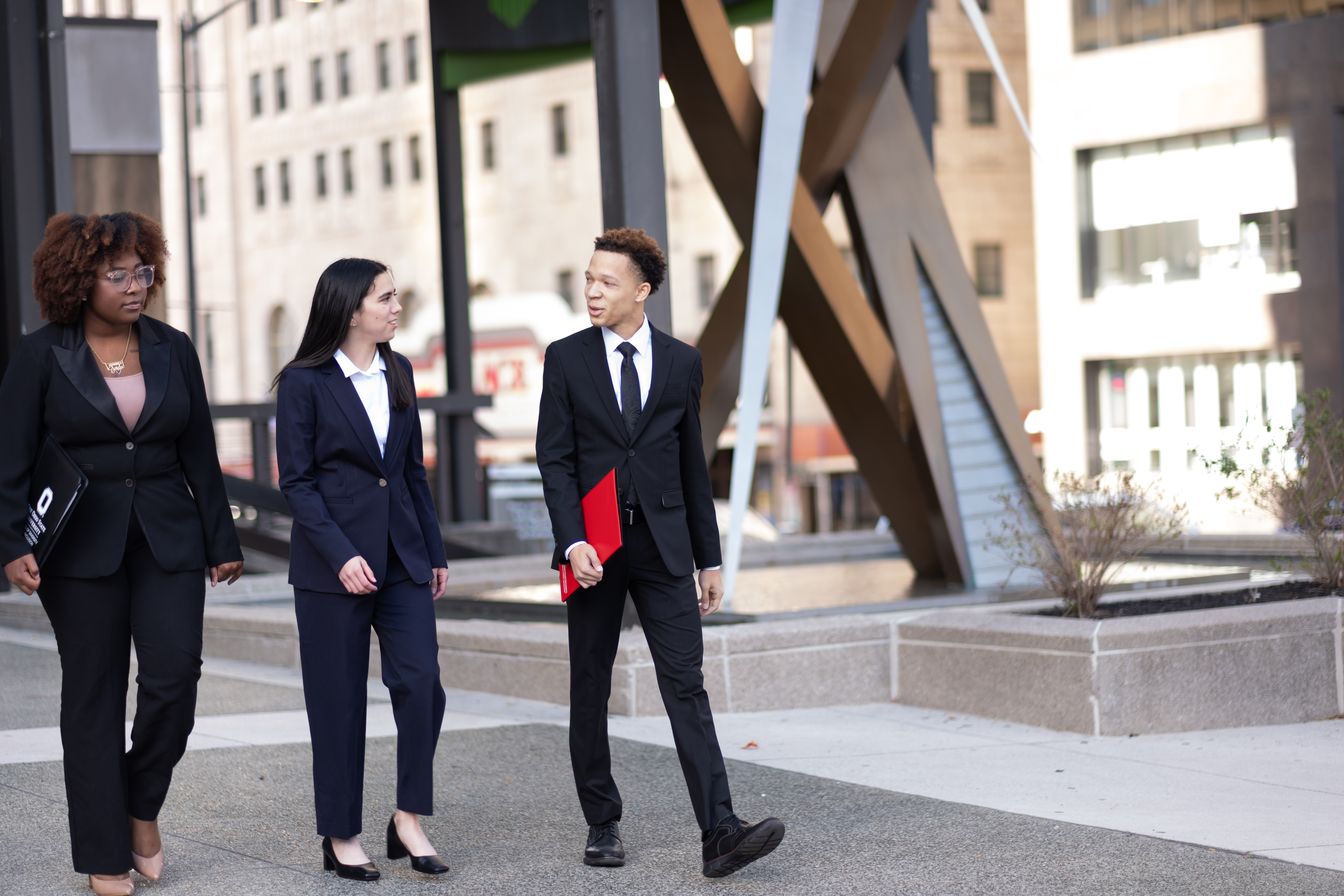 Three students in business attire walk and talk outdoors on campus, one carrying a red folder.Provide your feedback on BizChat