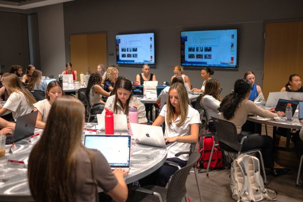 Students sitting at round tables working on their computers; screens in background projecting a power point presentation