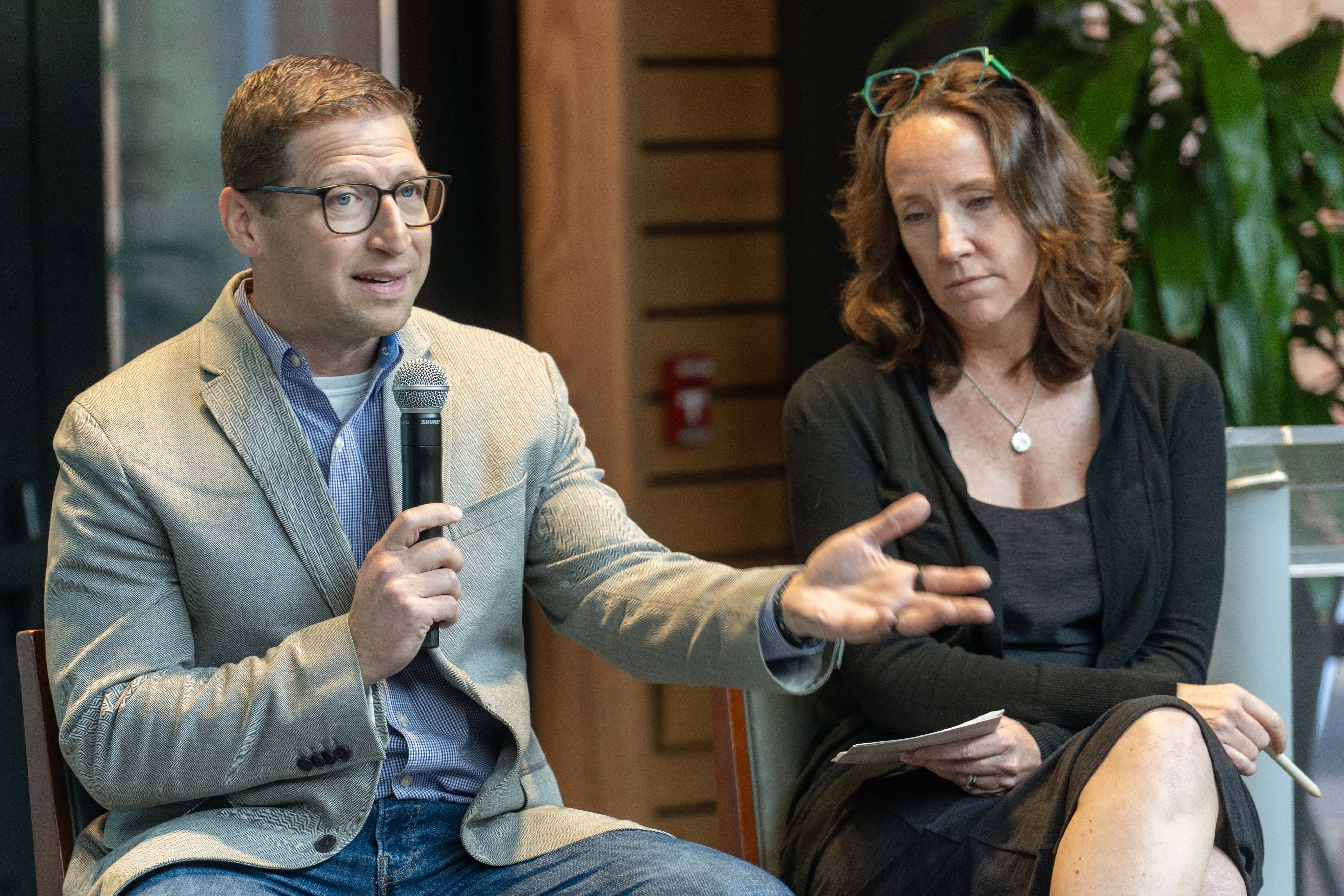 Dan Haber in a suit jacket and jeans is seated on a high stool and speaking into a microphone next to Joan Smith.