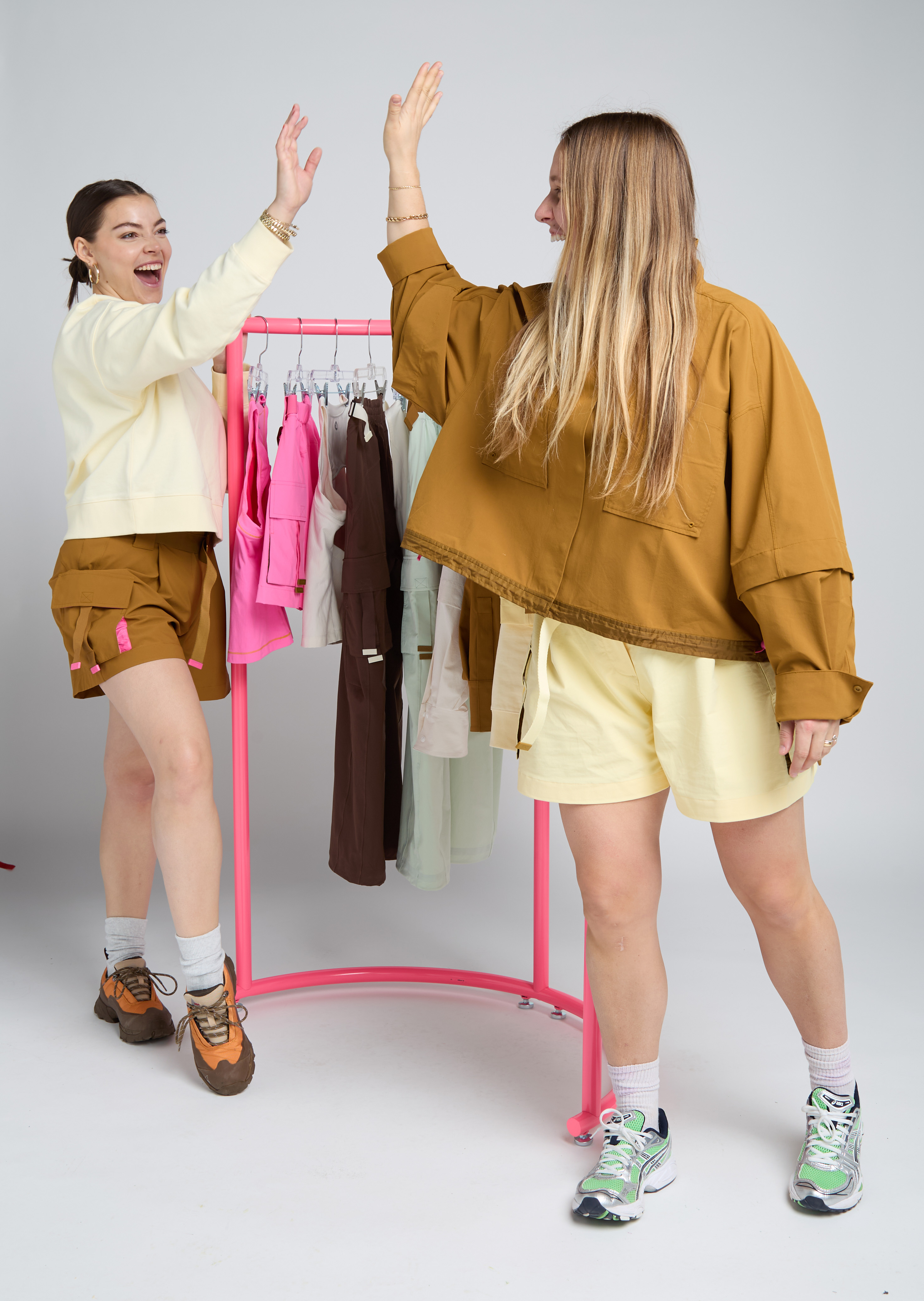 Madison Hilson and Valentina Thompson are raising their hands in a high five in front of a rack of outdoor clothing.