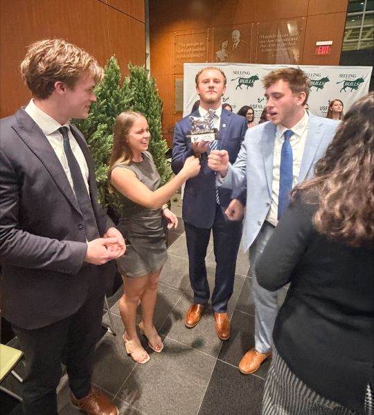 Four students in business attire fist bumping while showing off a trophy