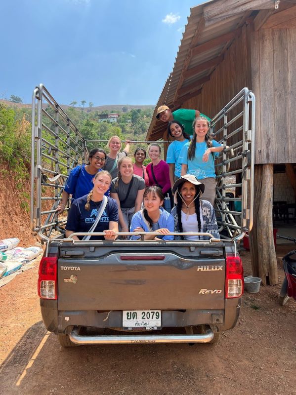 Group of people in the back of a pickup on a dirt road next to a wood building