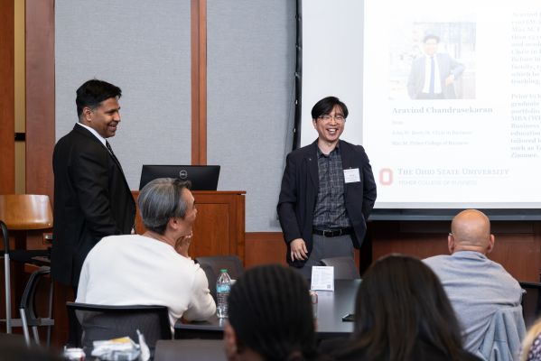Two men smiling in front of room with screen in background, people in foreground.