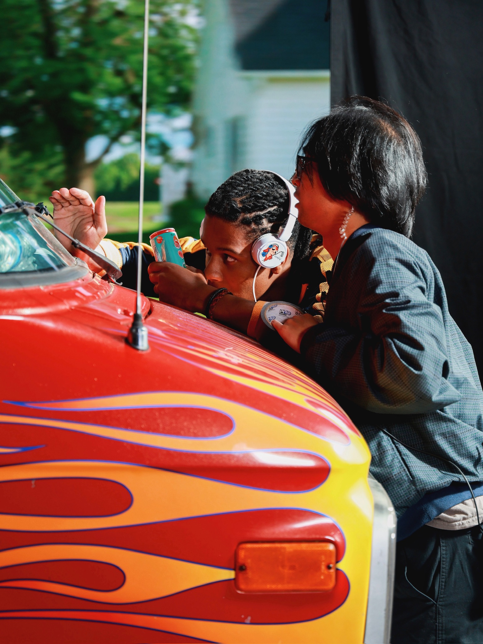 Justen Turner, left, and Enrique Cabotage, stand at the front of a red van with flames painted on it, checking a camera angle during filming. 