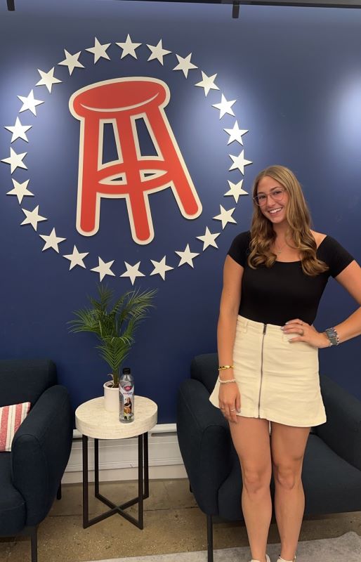 Woman in short skirt and short sleeved shirt stands in front of a logo of a barstool