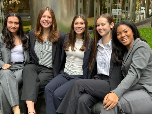 Five women in suites sitting outside with arms linked around each other