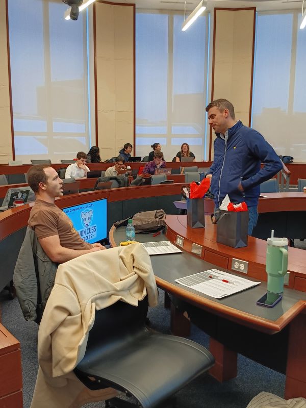 Man seated talking with man standing in semi-circle classroom with students in background
