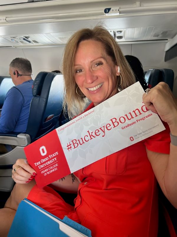 Woman in red dress sitting in an airplane seat holding a sign that says #BuckeyeBound